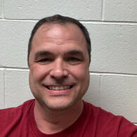 A smiling man with dark hair, wearing a red t-shirt